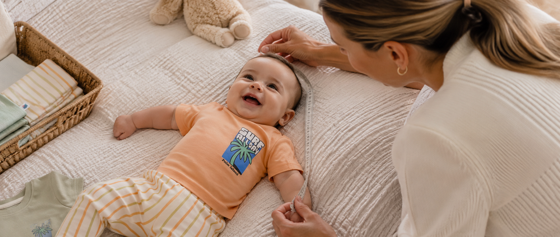 How to pick the right size for baby clothes – mother measuring a baby outfit with a measuring tape while baby boy smiles in a nursery.